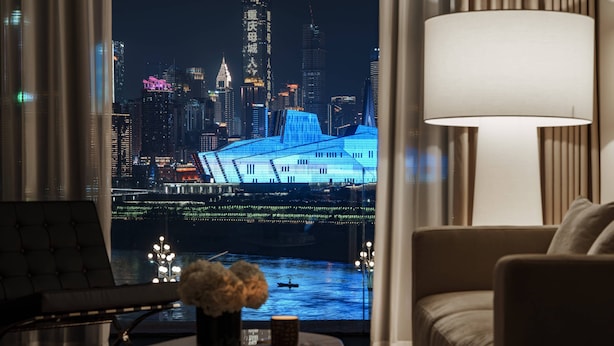 Una habitación con vistas a un horizonte urbano nocturno, un ventanal grande, un sofá, una silla y una mesa con un jarrón y un libro.