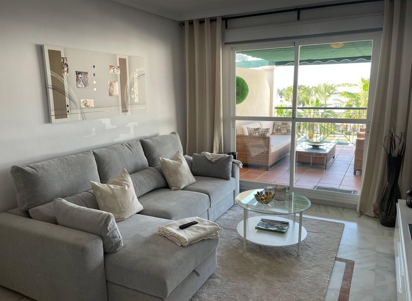 A modern living room with a grey sofa, a glass coffee table, and a view of an outdoor patio with furniture.