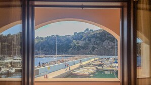 A view through a window of a marina with boats docked, people walking, and a hilly landscape in the background. - Casa del porticciolo di Capraia (Porto)