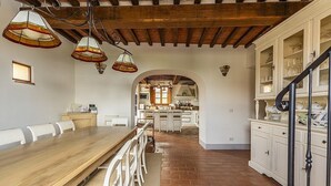 Une vaste salle à manger avec une longue table en bois et des chaises blanches, une cuisine à l’arrière-plan et un escalier avec une armoire blanche sur le côté droit.