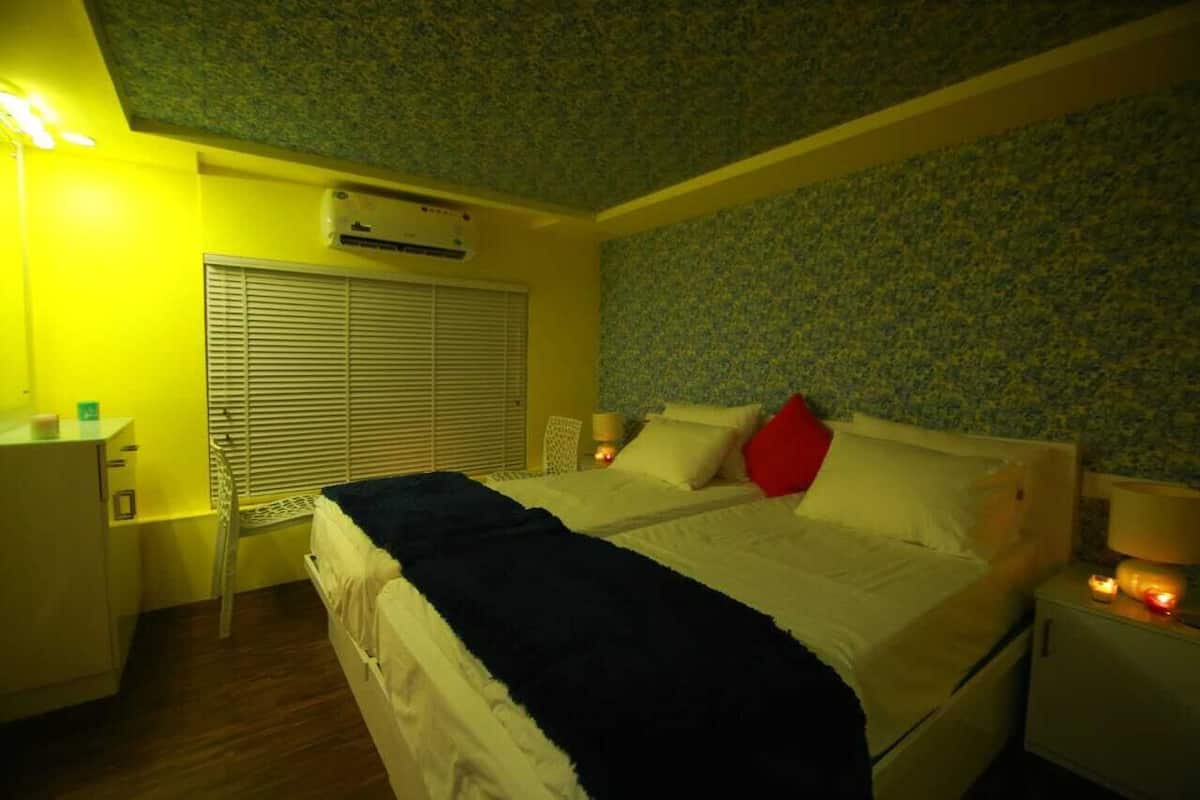 A bedroom with a bed, bedside tables, a window with blinds, an air conditioning unit, and a wall with a floral pattern.