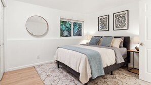 Una camera da letto con un letto, uno specchio rotondo, due quadri incorniciati, una finestra vista alberi e una porta.