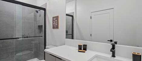 A modern bathroom with a glass shower enclosure, a toilet, a large mirror, and a white countertop with a sink and two black soap dispensers.