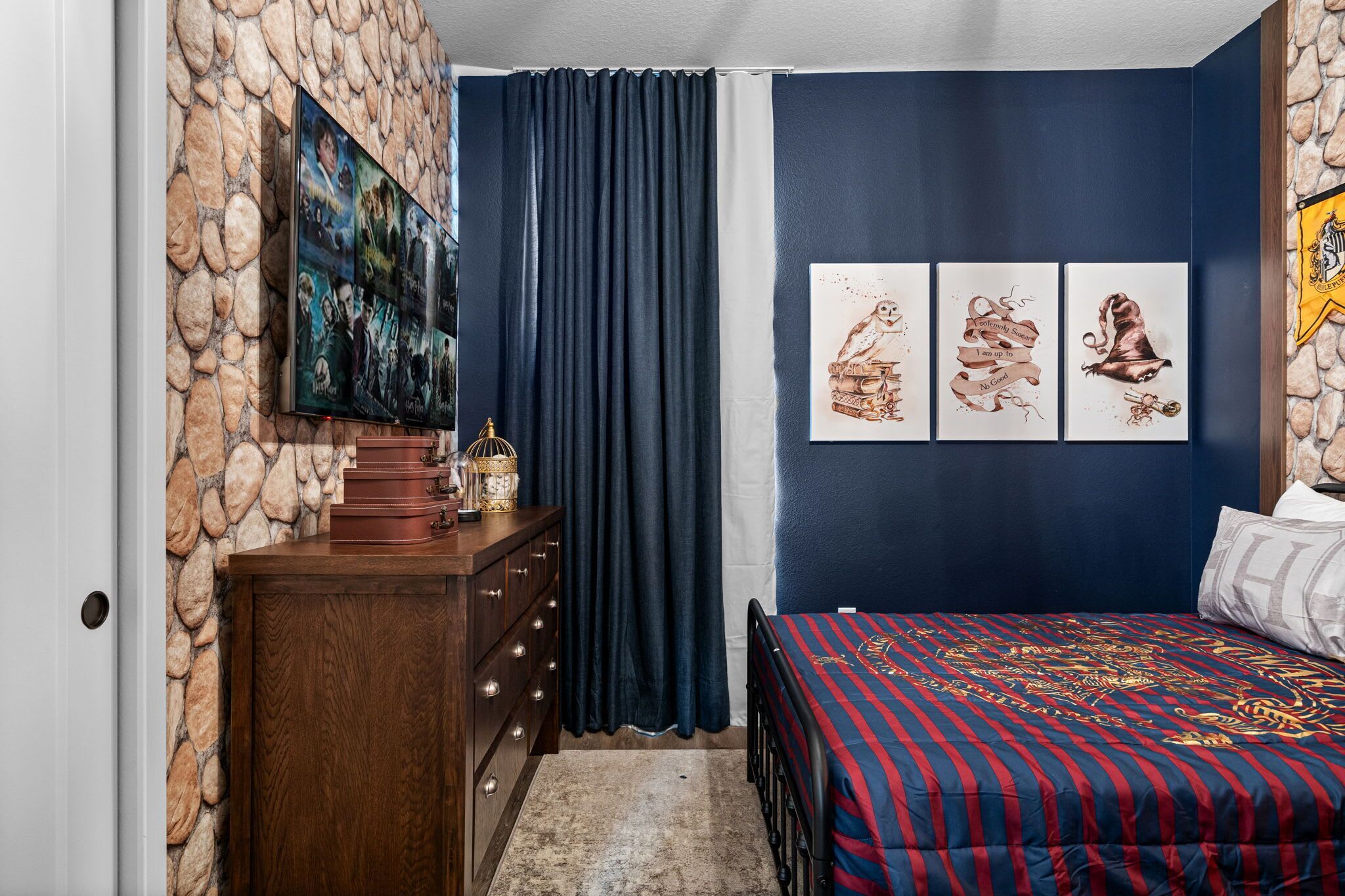 A bedroom with a stone wall, a large TV, a bed with a striped blanket, and three owl-themed pictures on the wall.