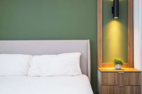 A neatly made bed with white linens, a bedside table with a small plant, and a wall-mounted light fixture.