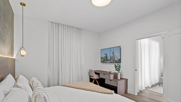 A modern hotel room with a large bed, a desk, and a chair. There is a painting on the wall and a window with curtains.