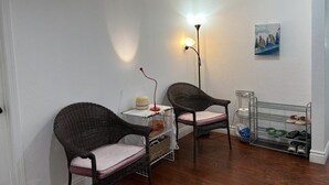 Une petite pièce avec deux fauteuils en osier, une lampe sur pied, un tableau accroché au mur et un porte-chaussures.