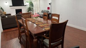 Un coin repas avec une table en bois, six chaises et un centre de table floral. Un canapé et un téléviseur se trouvent à l’arrière-plan.