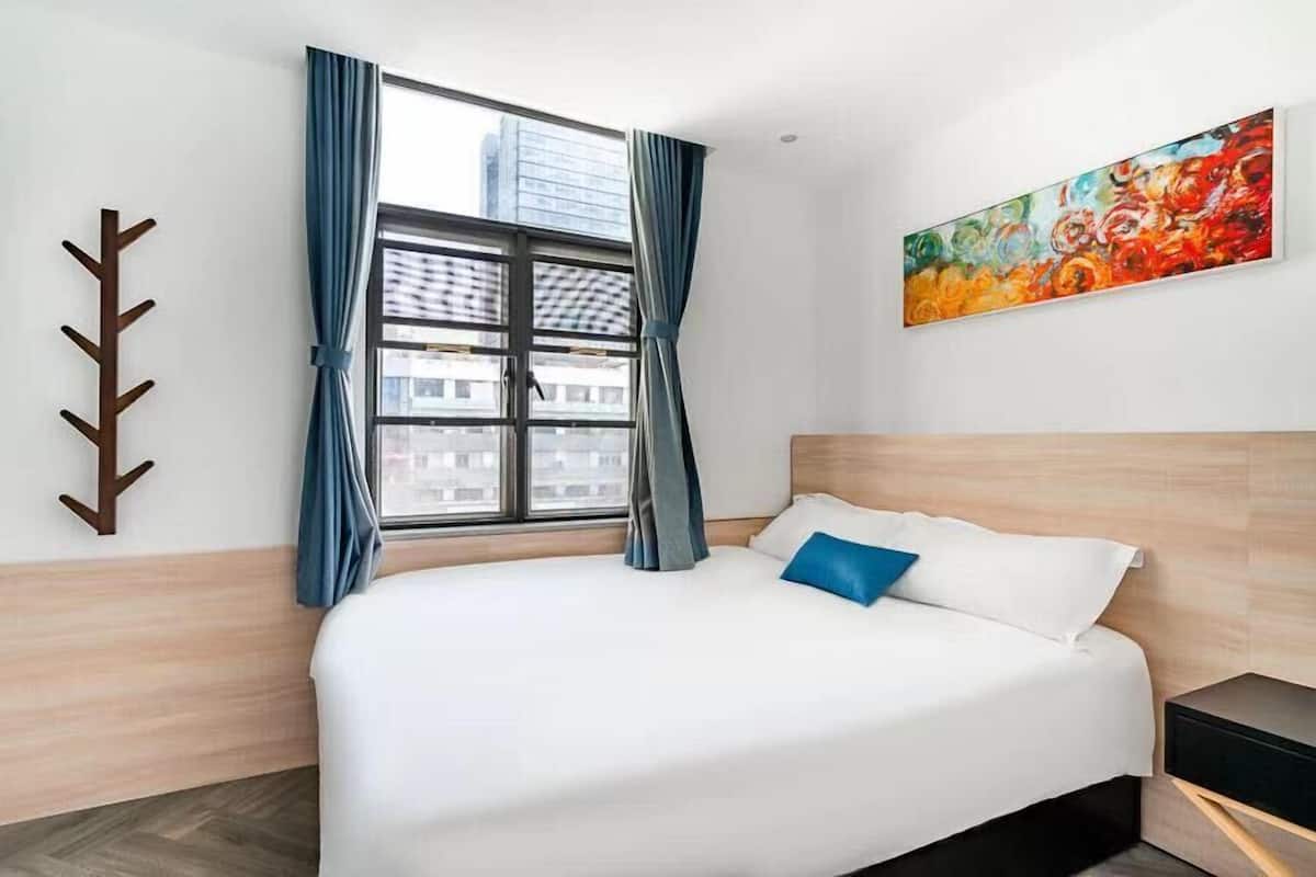 A hotel room with a large bed, a window with curtains, a painting on the wall, and a coat rack.