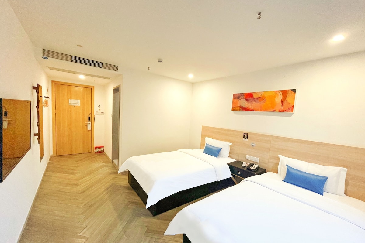 A hotel room with two beds, a wooden headboard, a painting on the wall, and a hallway leading to another room.