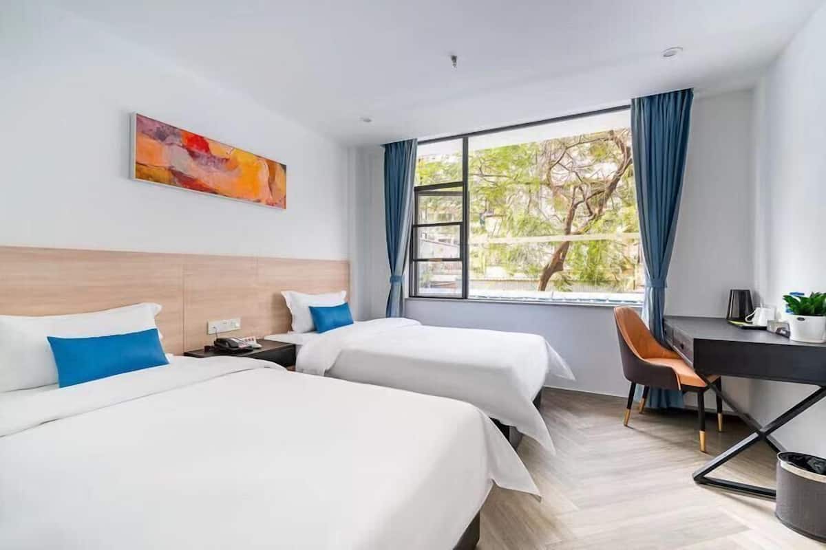 A hotel room with two beds, a desk, and a chair. There is a window with curtains, a painting on the wall, and a view of trees outside.