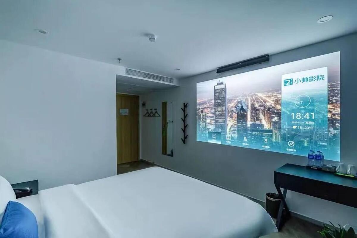 A hotel room with a bed, a desk, and a large digital display on the wall showing a cityscape and room temperature.