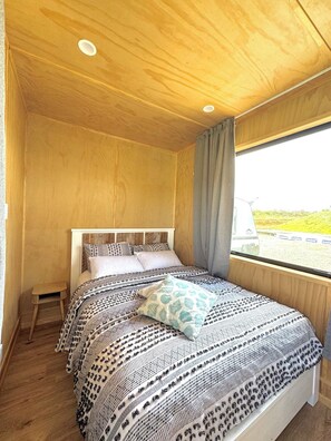A bedroom with a wooden ceiling, a bed with patterned bedding, a small wooden stool, and a window with a view of the outside. - Ataahua HighlandA little slice of Paradise- Off Grid retreat. (Te Kopuru)