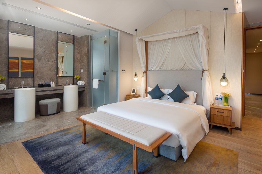 A modern hotel room with a large bed, a bedside table, a vanity with a mirror, and a shower area.