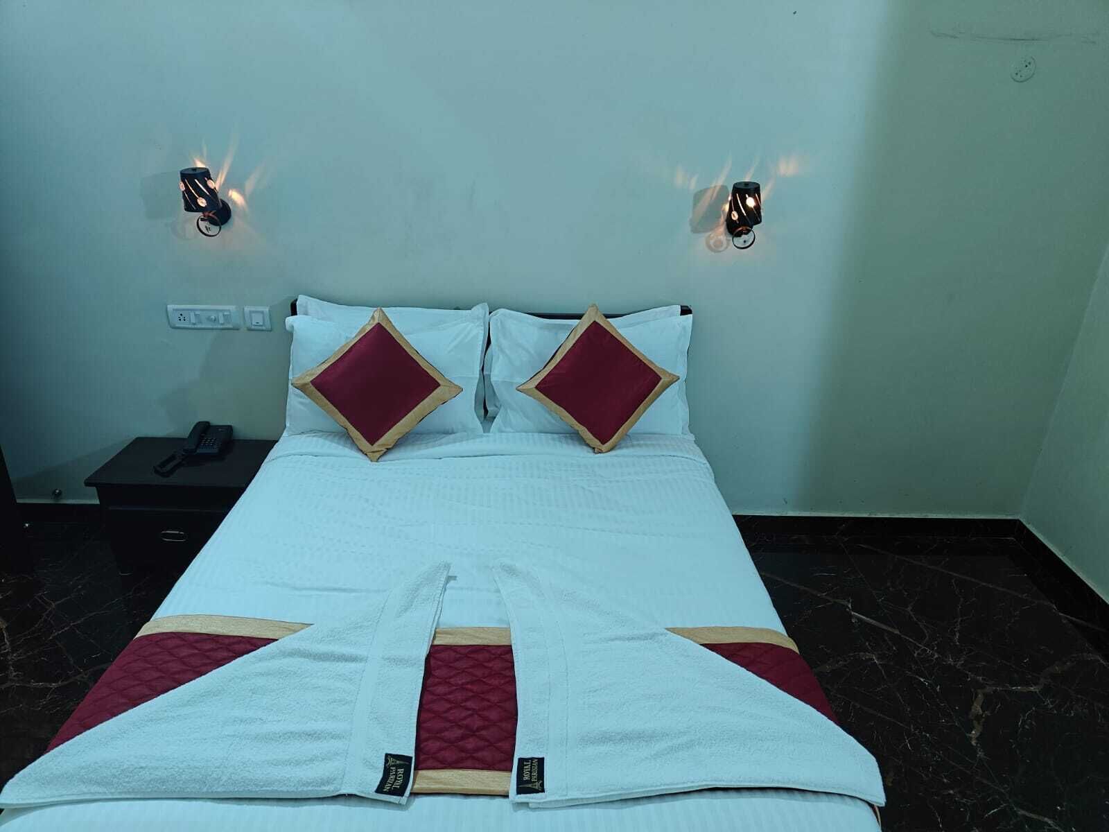 A hotel room with a bed, two red and gold pillows, a white bedspread, a white blanket, a black nightstand with a phone, and two wall-mounted light fixtures.