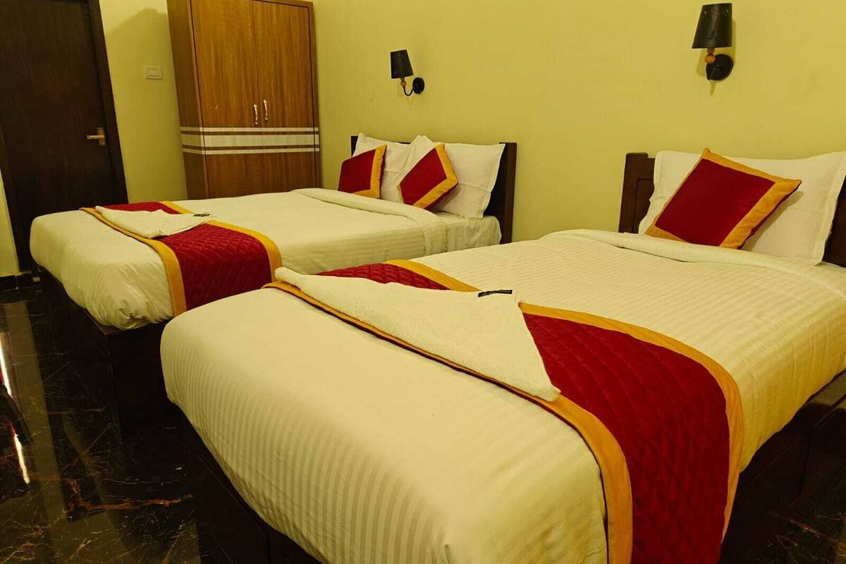 A hotel room with two beds, each with white bedding and red accents, a wooden wardrobe, and a wall-mounted lamp.