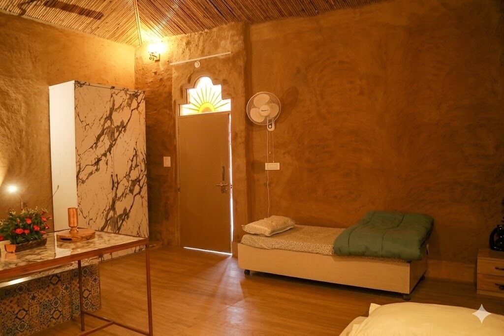 Une pièce avec un lit, un miroir, un ventilateur et une porte donnant sur une fenêtre ensoleillée.