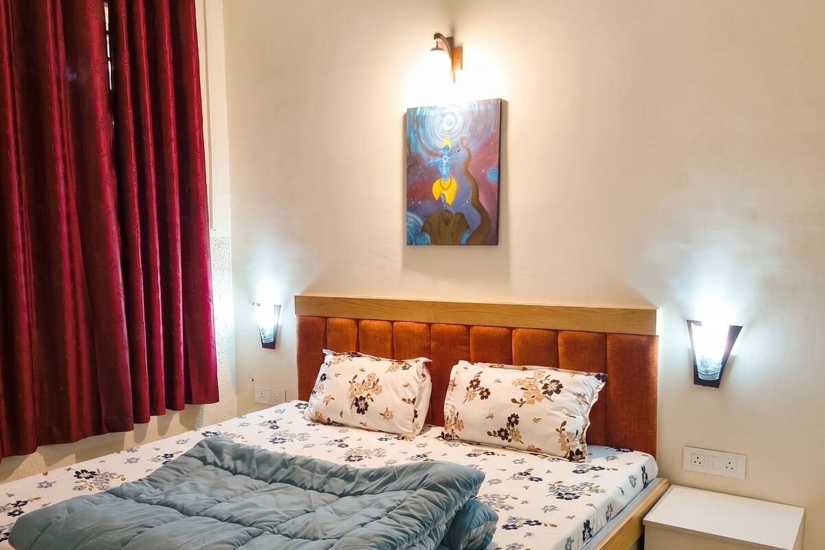 Quarto com cama, mesa de cabeceira, luminária na parede, ventilador de teto e um quadro.