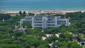 Un edificio moderno con techo plano, rodeado de árboles verdes frondosos, con vista a una playa con sombrillas y gente.