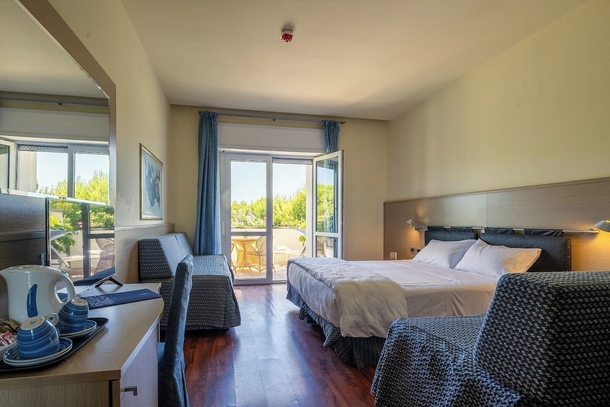 Habitación de hotel con una cama, un escritorio con una computadora y una tetera, una silla y vistas al exterior a través de un ventanal.