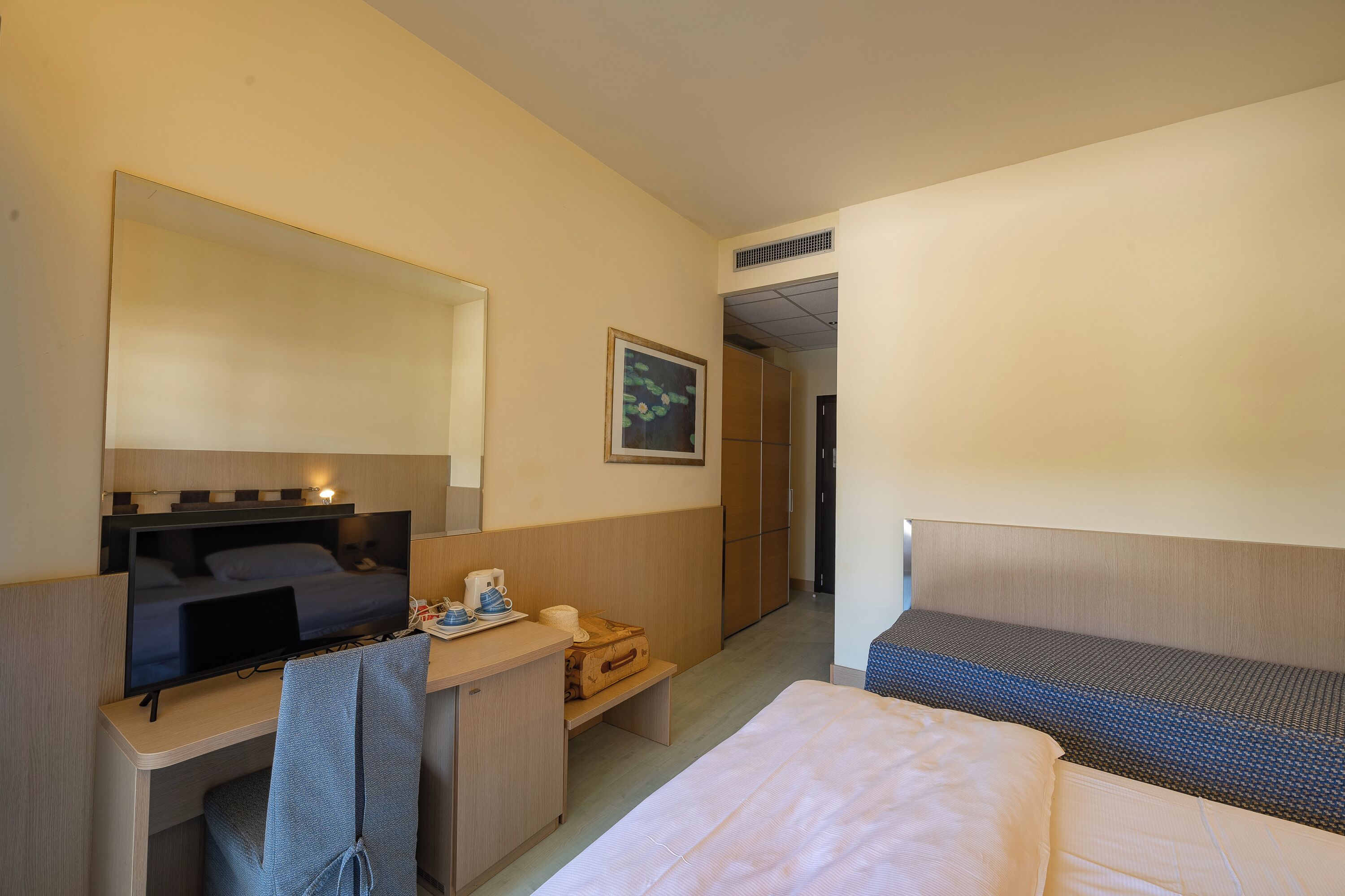 A hotel room with a bed, a desk with a computer, a mirror, a small table with a cup and a suitcase, and a door leading to another room.