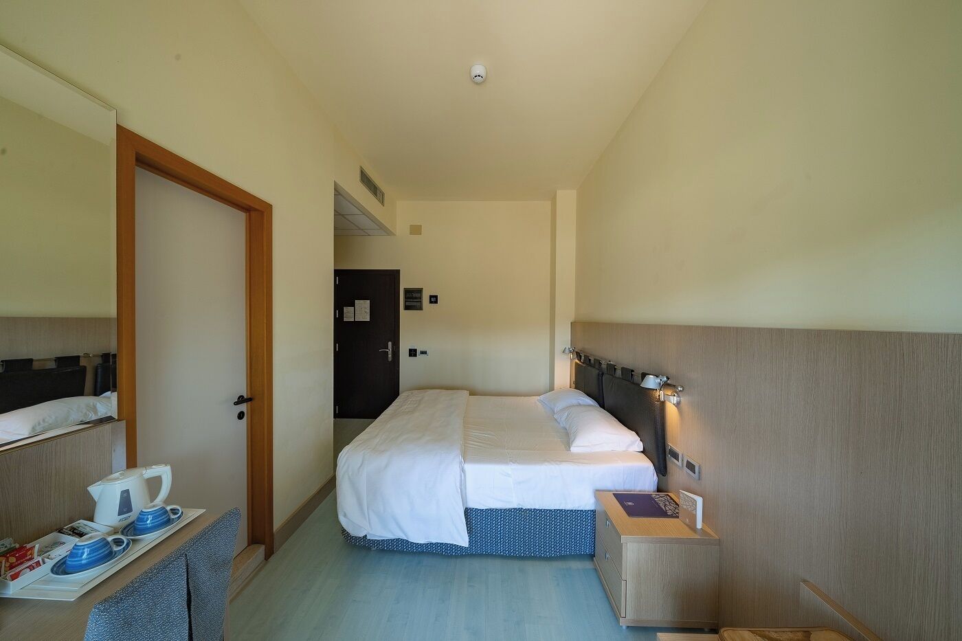 Hotellihuone, jossa on parivuode, yöpöydät, pieni työpöytä, jossa on vedenkeitin ja kuppeja, sekä ovi, joka johtaa toiseen huoneeseen.