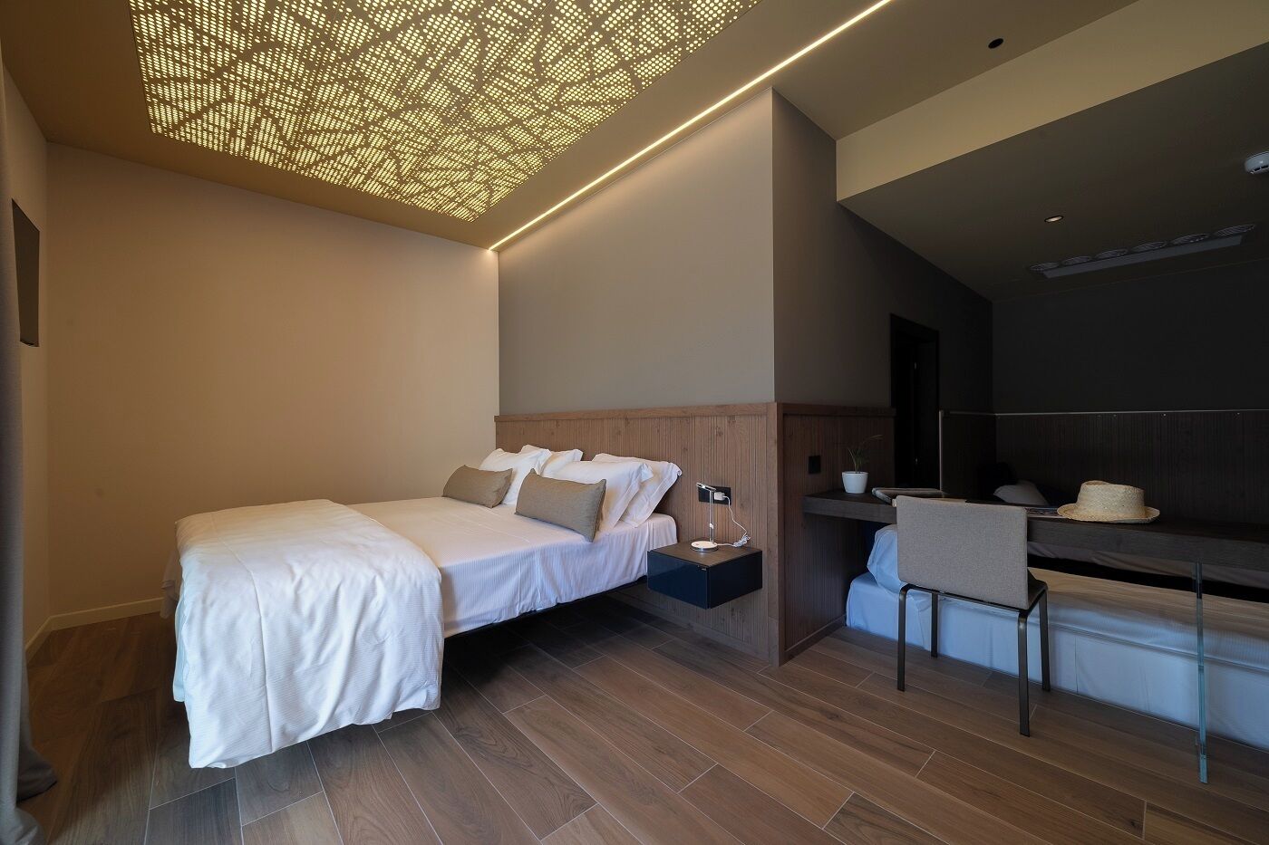 A modern hotel room with a bed, bedside table, lamp, and a small seating area with a chair and a hat.