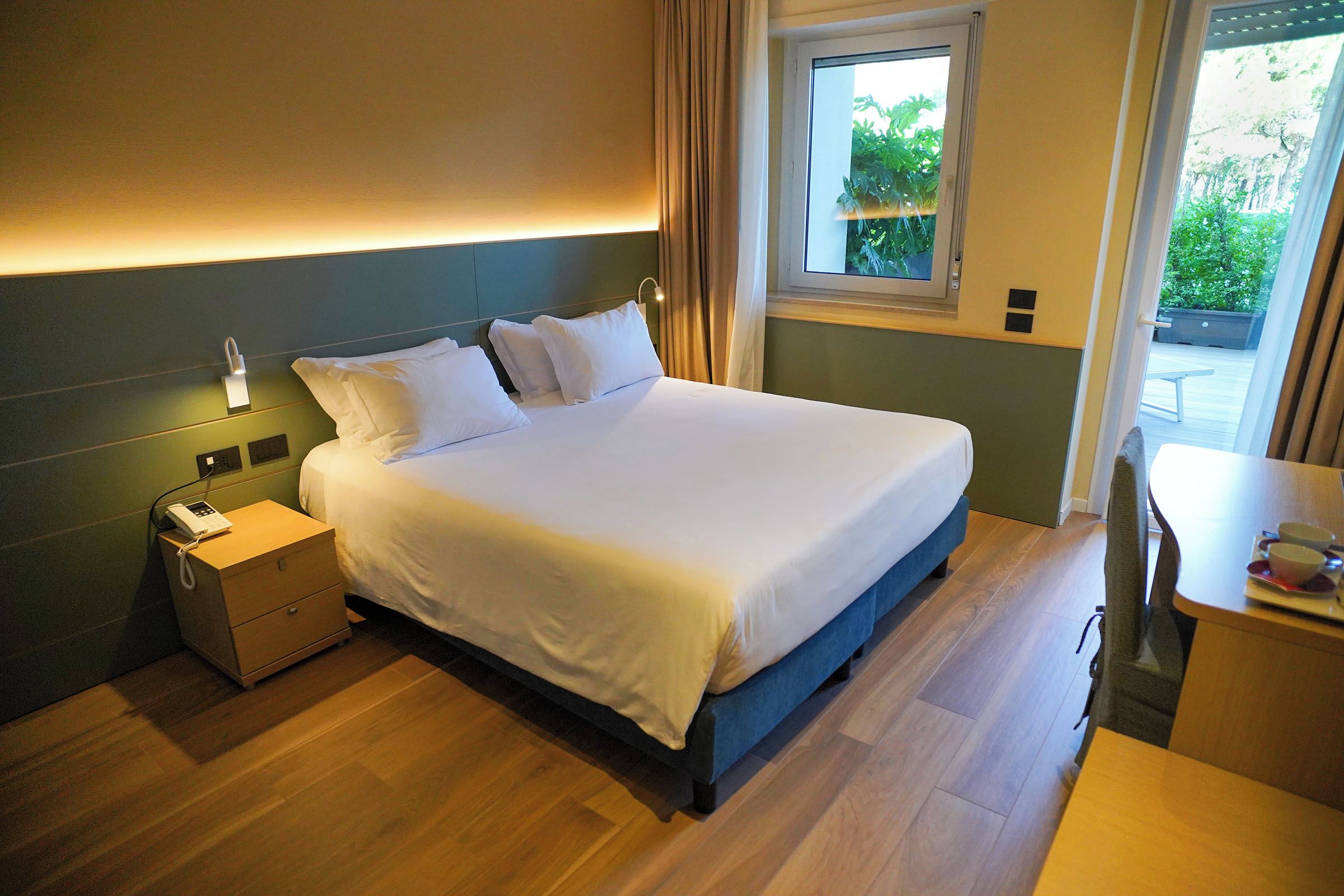 A hotel room with a bed, bedside table, lamp, and a window with a view of greenery.