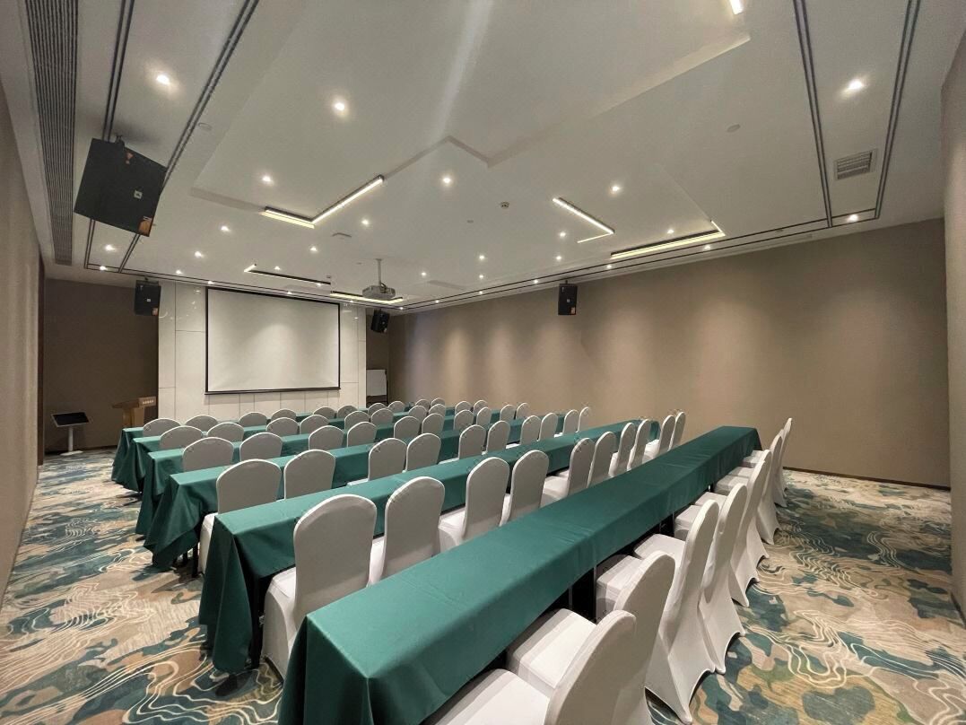 A conference room with rows of tables and chairs, a projector screen, and a carpeted floor.