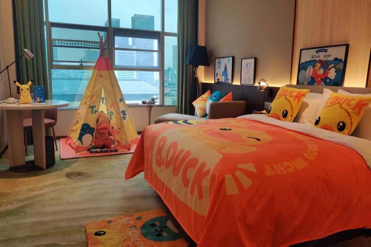 A hotel room with a large bed featuring a Duck-themed bedspread, a small table with a lamp, a chair, a desk with a computer, a window with a city view, and a tent with a cartoon character.