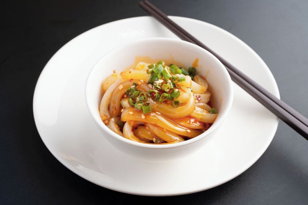 Una ciotola di noodles guarniti con scalogno verde e semi di sesamo su un piatto bianco.