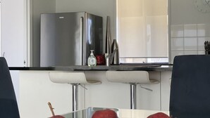 A modern kitchen with a glass-top dining table, a stainless steel refrigerator, and a window with blinds.