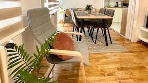 Een moderne woonkamer met een bank, eettafel en stoelen, een keuken op de achtergrond en een groot raam met jaloezieën.