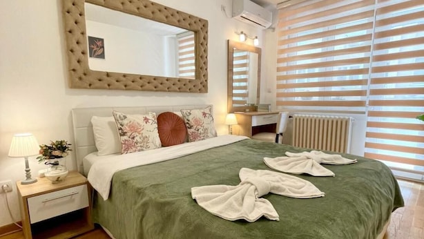 Een slaapkamer met een bed, nachtkastje, spiegel en raam met jaloezieën.