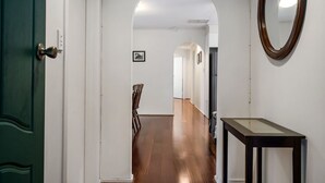Un couloir avec un plancher en bois, des murs blancs et une porte verte. Une petite table avec un miroir se trouve sur la droite.
