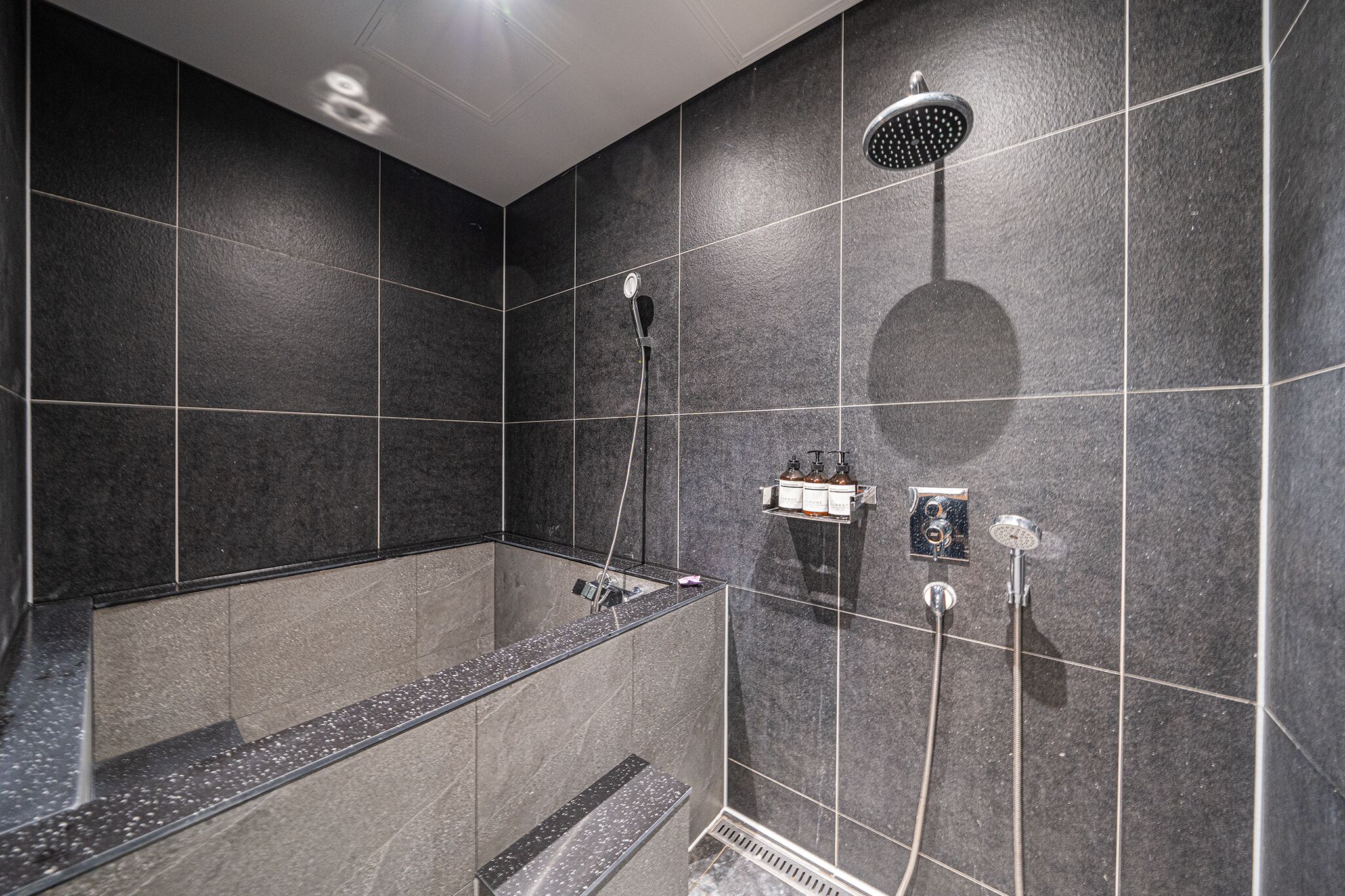 A modern bathroom with dark tiles, a glass-enclosed shower, and a built-in shelf with toiletries.