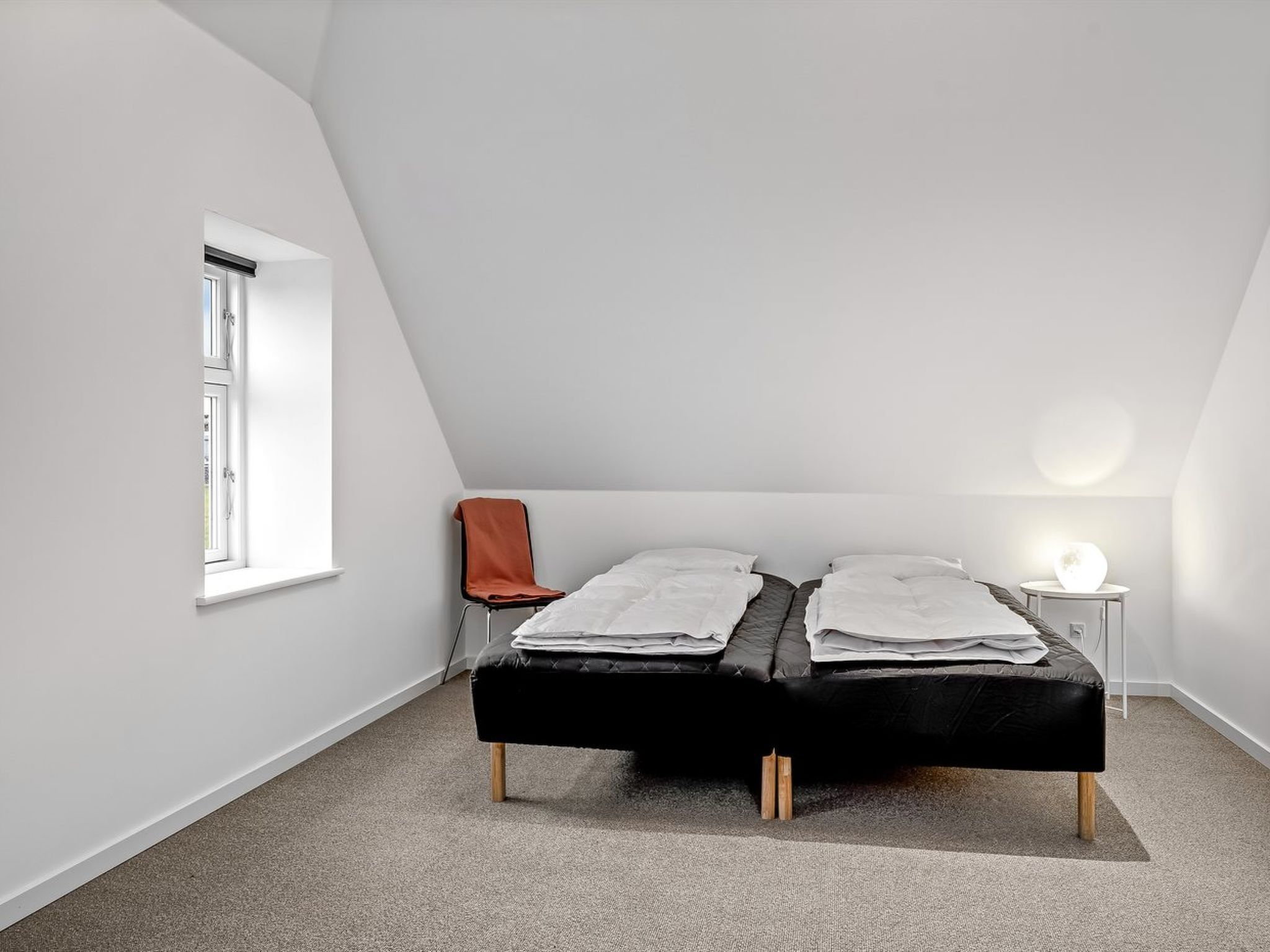 Een slaapkamer met een schuine kap, een enkel bed, een stoel, een klein tafeltje met een lamp en een raam.