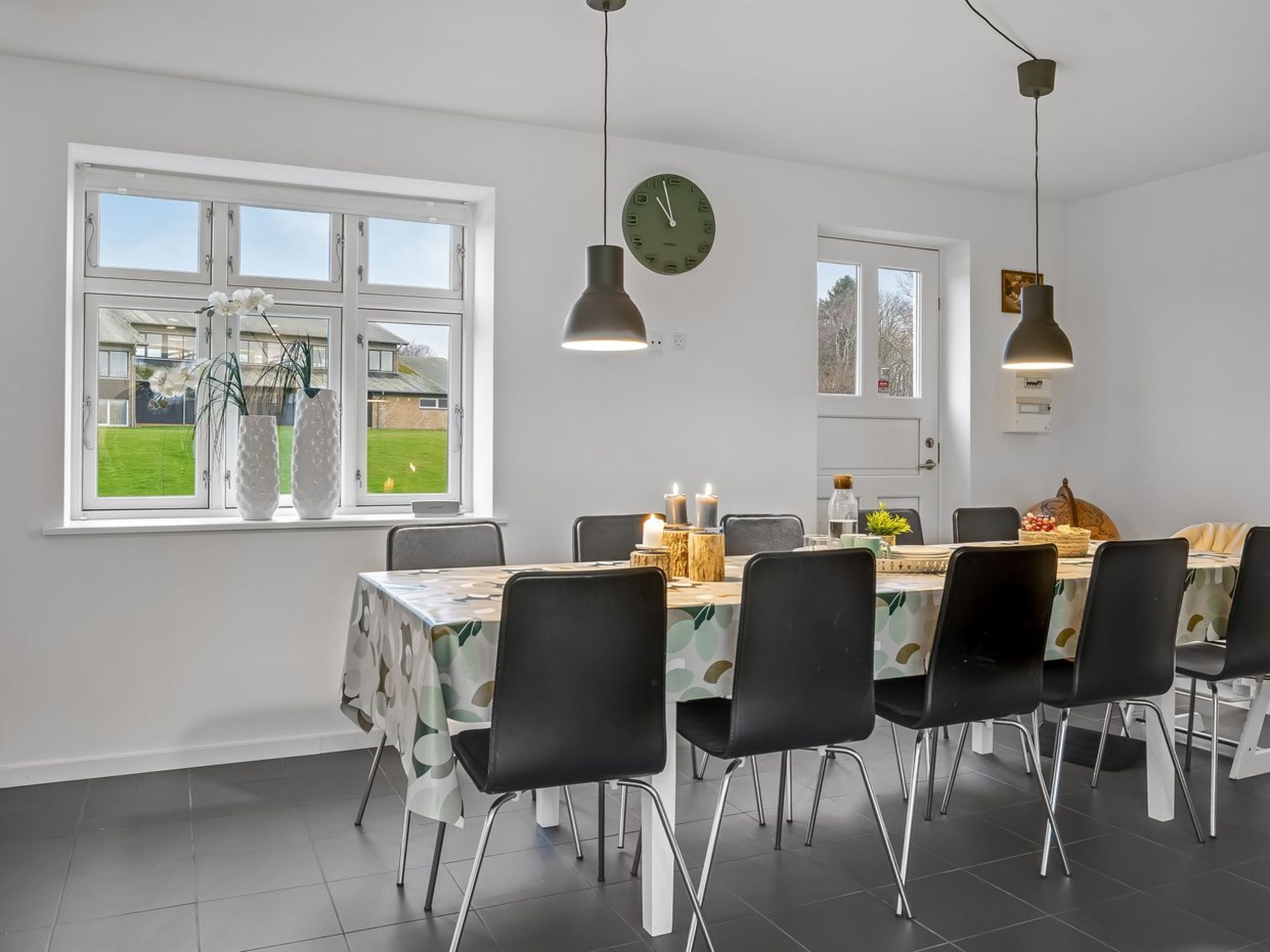 Een eetruimte met een tafel voor zes personen, omringd door zwarte stoelen. Boven de tafel hangen twee hanglampen en er is een klok aan de muur.