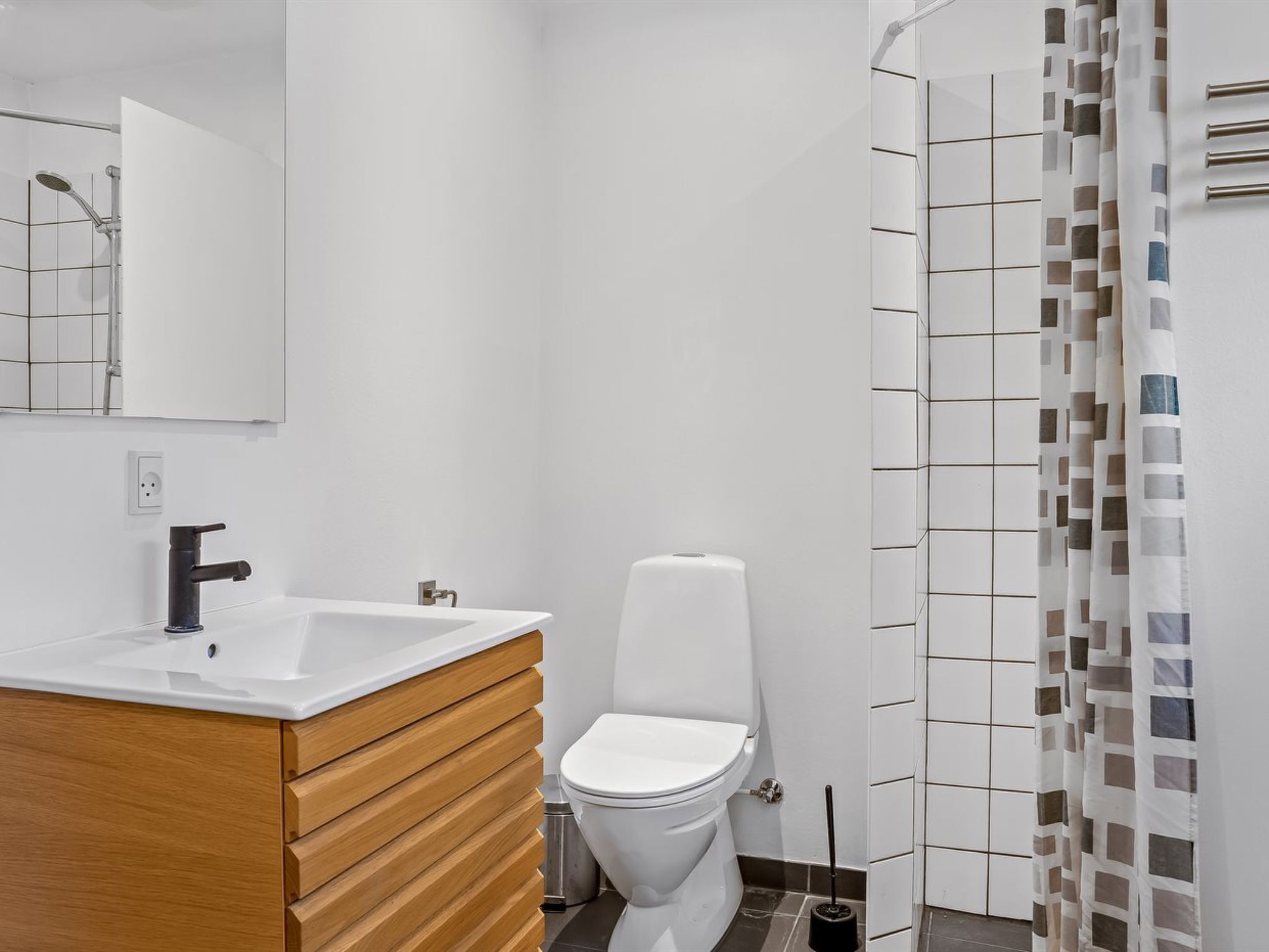Un baño moderno con lavamanos blanco, tocador de madera, inodoro y ducha con cortina estampada.