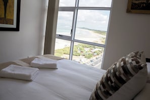 A hotel room with a large window offering a view of a beach and greenery.