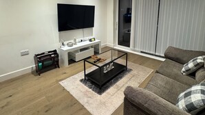 A modern living room with a flat-screen TV, a glass coffee table, a sofa, and a sliding glass door leading to a balcony. - Cosy Riverview apartment. 10mins to the o2 Arena. (Woolwich)