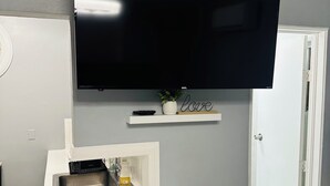 A modern living room with a flat-screen TV mounted on a wall, a small white shelf with decorative items, and a built-in sink. - Odriozola’s Vrbo Studio (El Paso)