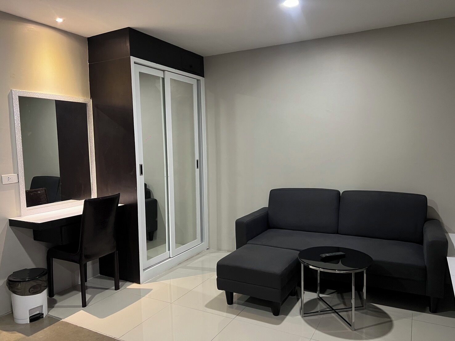 Una sala de estar moderna con un sofá negro, una mesita de centro redonda, una silla negra, un escritorio con espejo y un armario con puerta de vidrio corredera.
