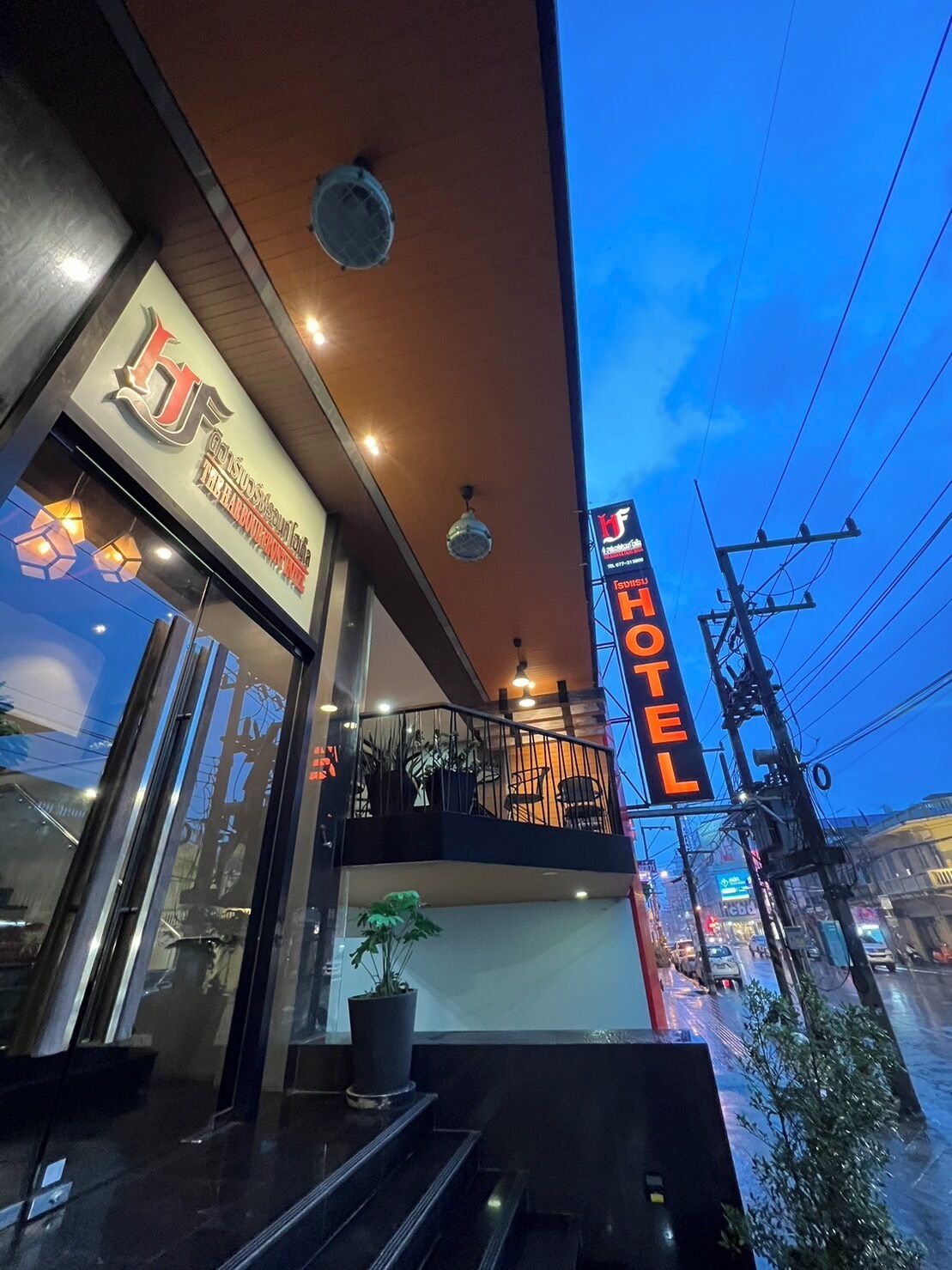 A hotel with a prominent 'HOTEL' sign, a balcony with chairs, and a potted plant on the ground floor.
