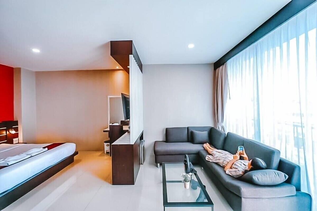 Una habitación de hotel moderna con cama, sofá, una mesa de centro de vidrio y un gran ventanal con cortinas.