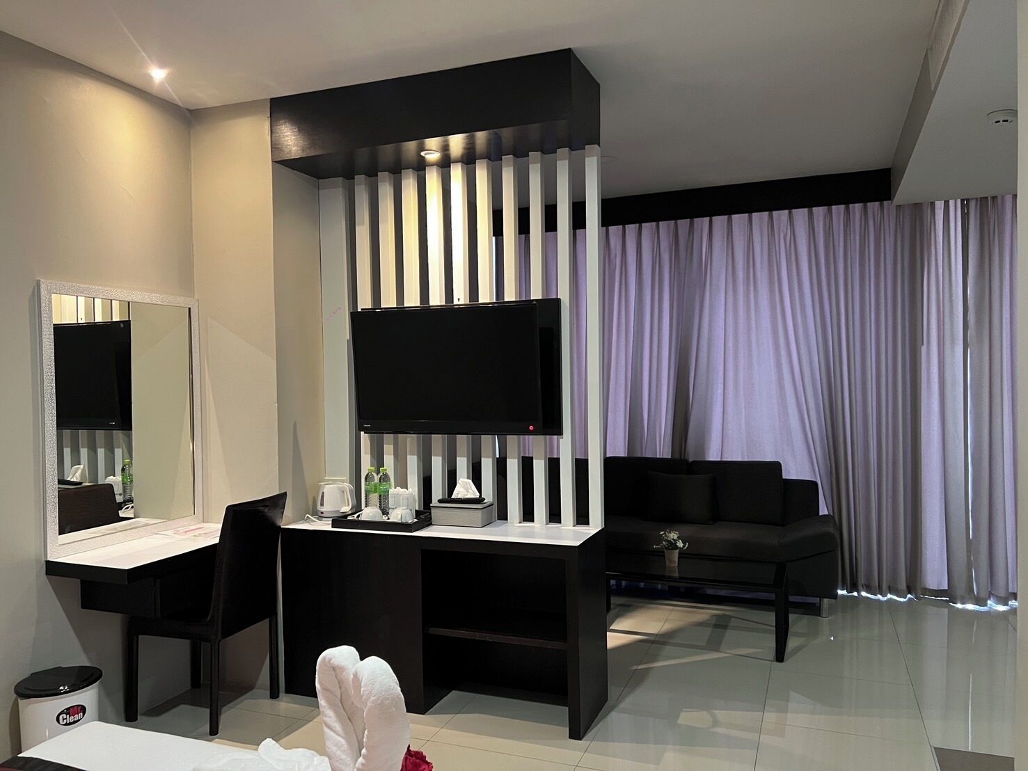 A modern hotel room with a large flat-screen TV, a desk with a chair, a sofa, and a large window with curtains.