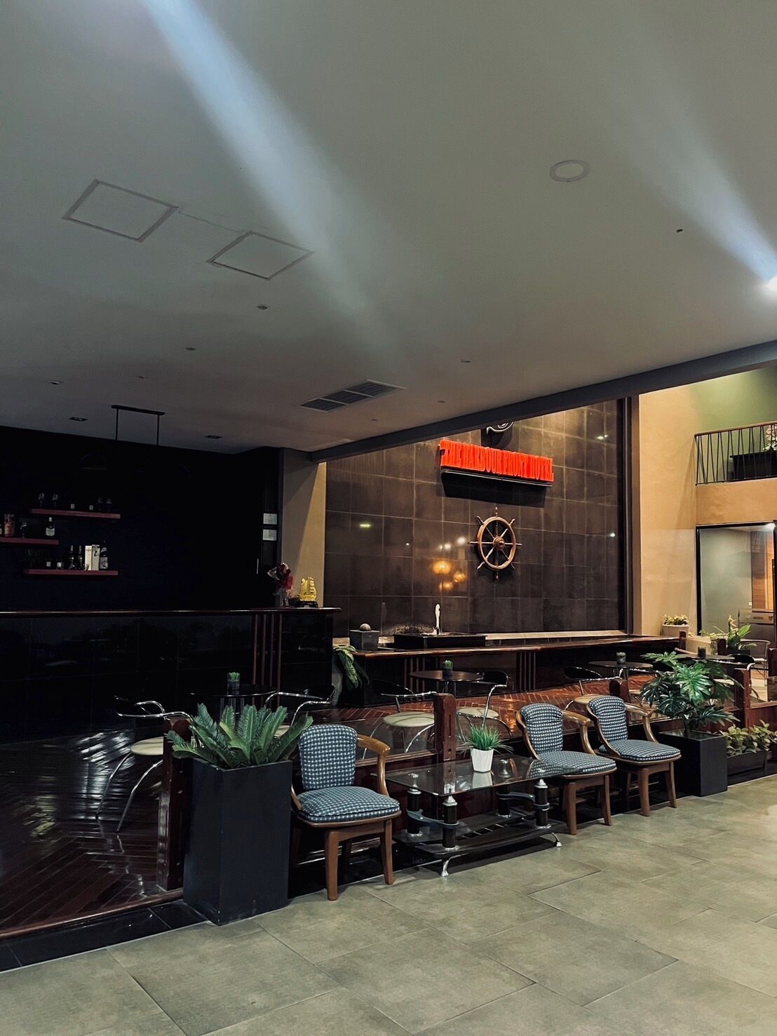 Zona de bar moderna con una gran decoración de timón de barco, asientos con plantas en macetas y un interior de tonos oscuros.