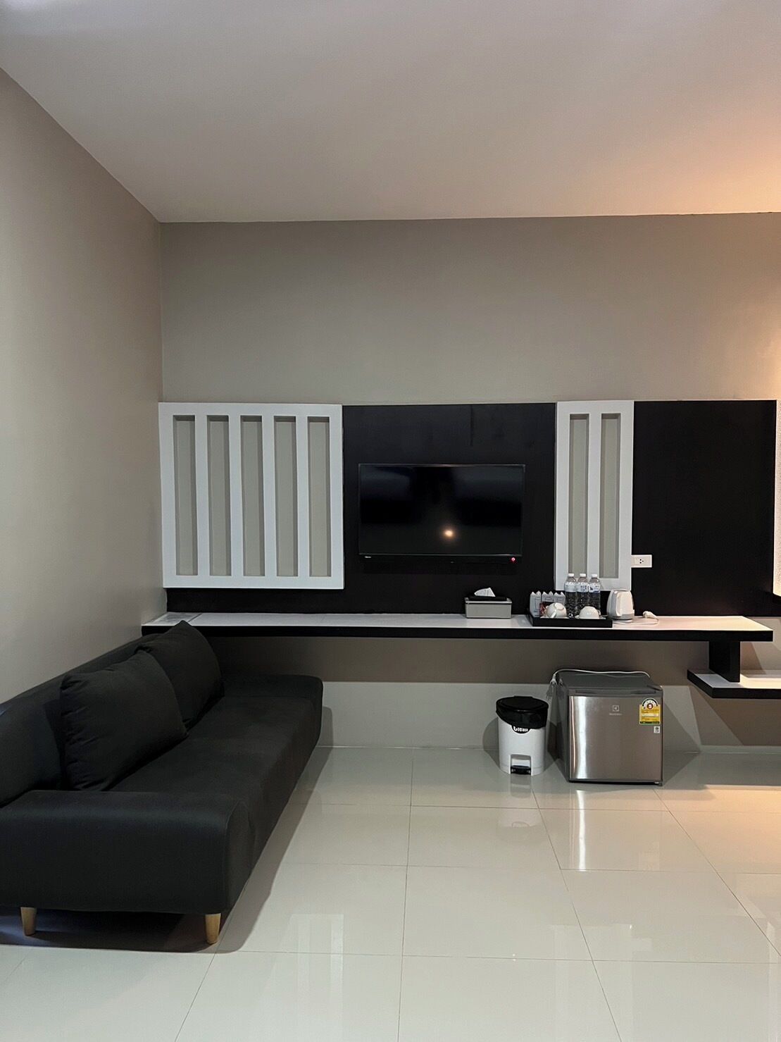 A modern living room with a black sofa, a flat-screen TV mounted on a black wall, and a small kitchen area with a coffee maker and a toaster.