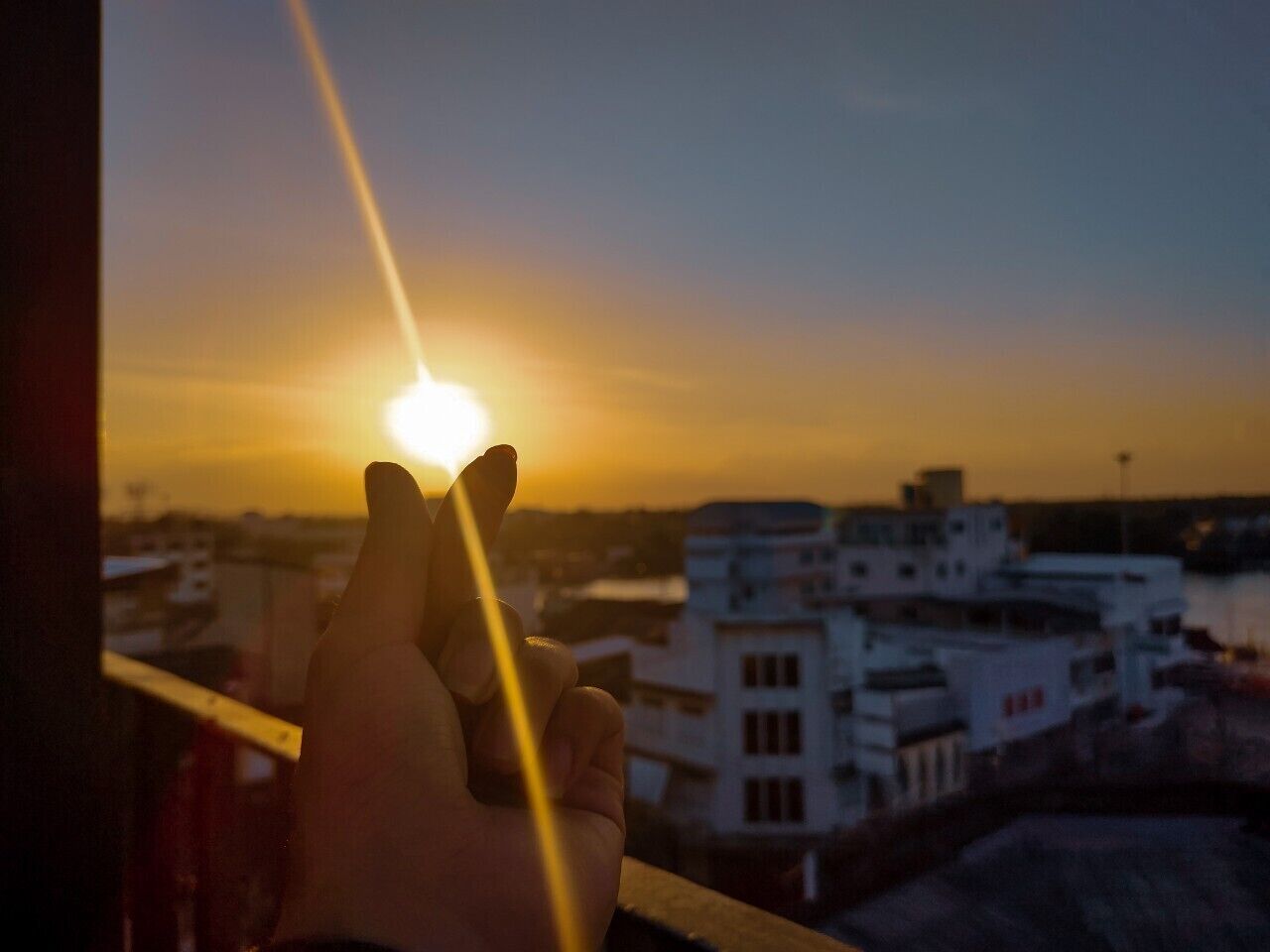 Una mano sostiene un vaso con vistas a un atardecer a través de una ventana, con vista a un paisaje urbano de edificios.