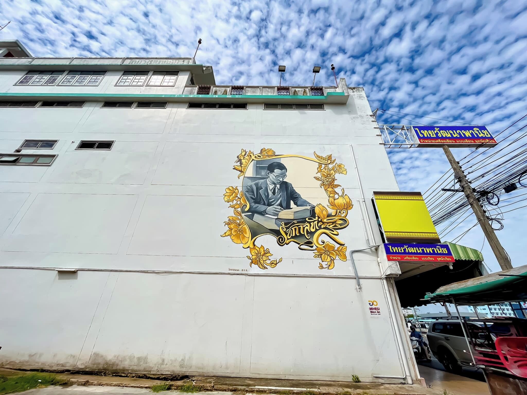 A building with a mural of a man writing, surrounded by ornate designs. There are signs in a foreign language, and a vehicle is parked nearby.